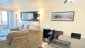 Ein Wohnzimmer mit beiger Couch, einem Esstisch mit Stühlen, einem Bücherregal und einem gerahmten Bild von einer Berglandschaft.