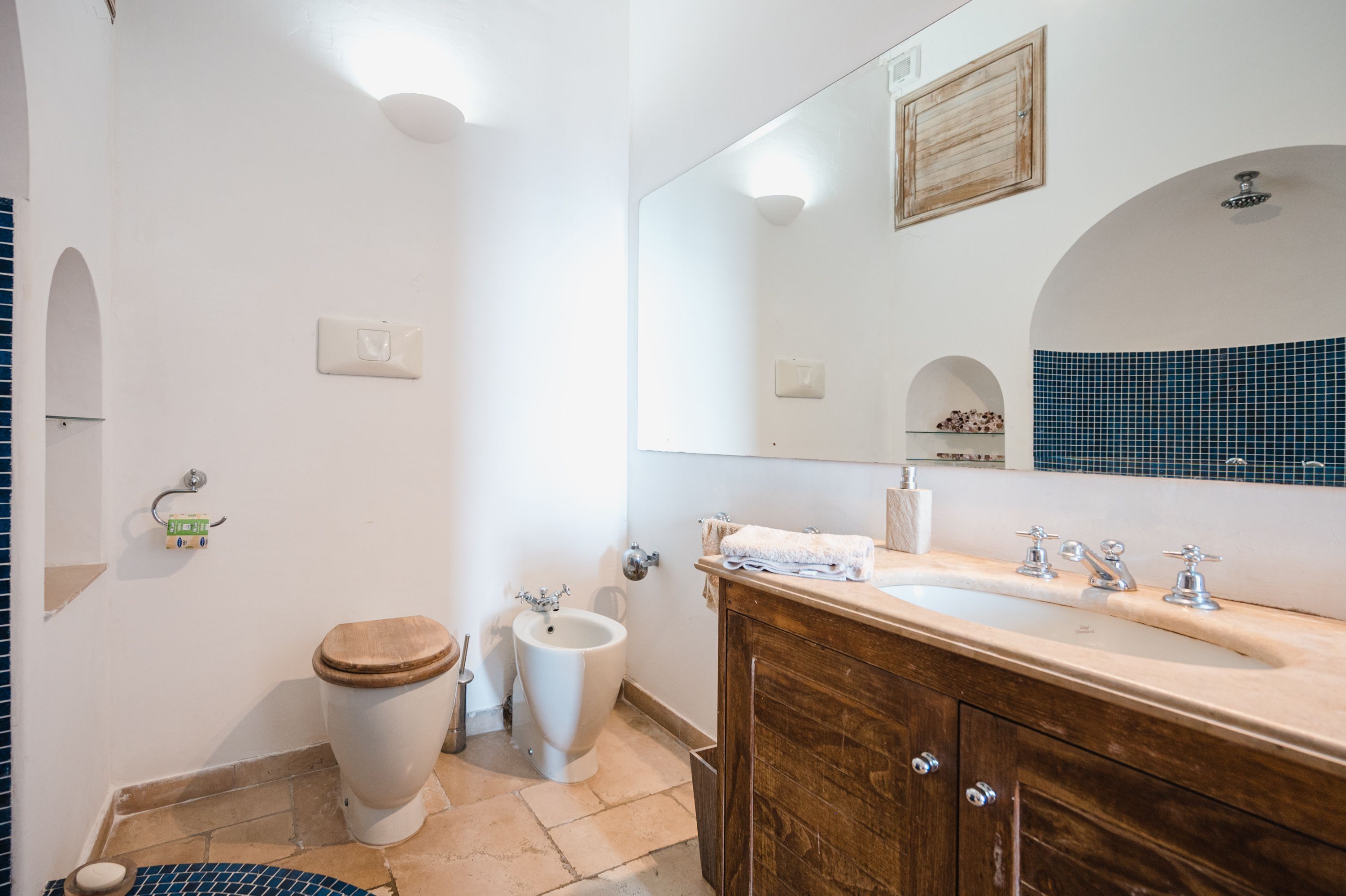 Un baño con inodoro de madera, un bidé, un lavabo con espejo y un salpicadero de azulejos.