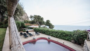 A swimming pool with lounge chairs and an umbrella, surrounded by greenery and overlooking a body of water. - Villa Michika by Elite Villas (Capri)