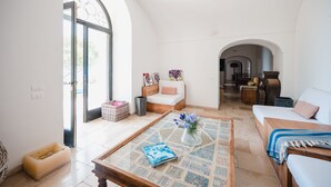 A spacious living room with a large arched window, a sofa, a coffee table with a book and vase, and a wooden bench. - Villa Michika by Elite Villas (Capri)