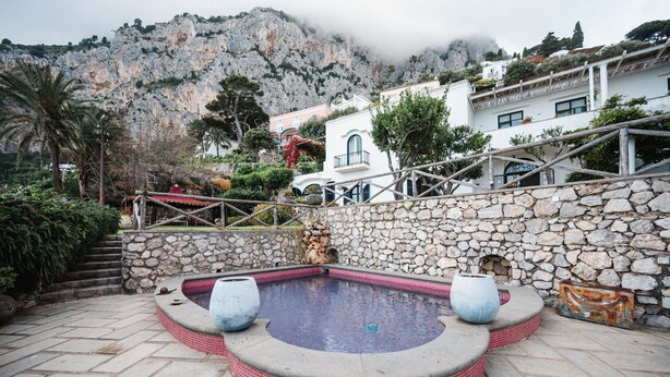 A stone pool with steps leading up to a white building with a balcony, surrounded by greenery and a mountainous backdrop. - Villa Michika by Elite Villas (Capri)