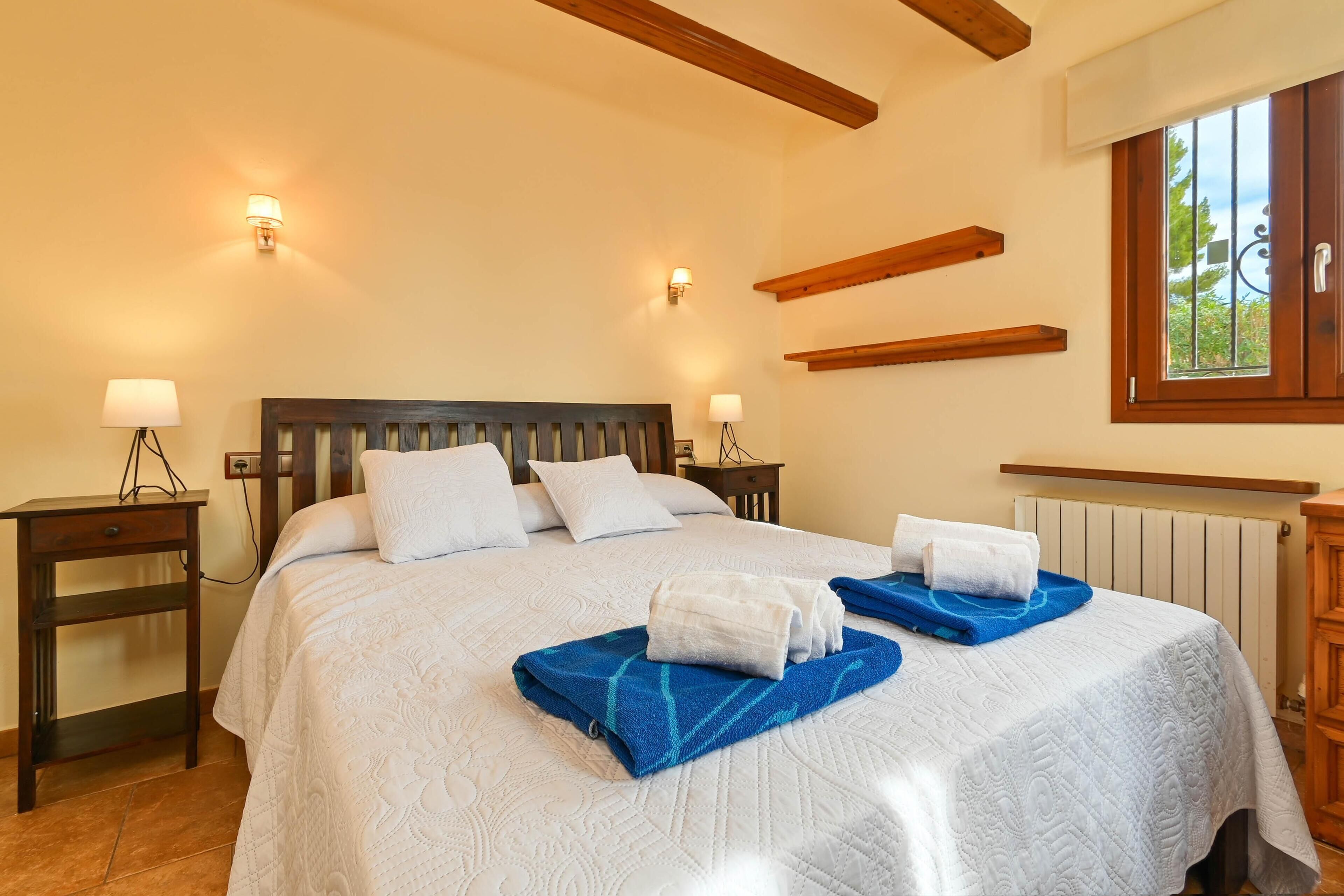 A well-lit bedroom with a large bed, two bedside tables, and a window with a view of greenery.