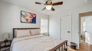 Un dormitorio con cama, ventilador de techo, un cuadro, mesita de noche, lámpara, ventana con persianas y puerta que da a otra habitación.