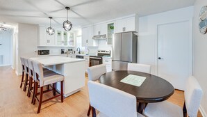 Una cocina moderna con armarios blancos, una isla central con taburetes y una mesa de comedor con sillas, además de un refrigerador.