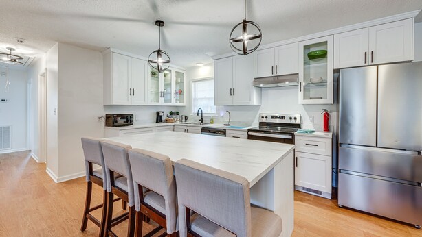 Una cocina moderna con armarios blancos, una isla grande con taburetes de bar, electrodomésticos de acero inoxidable e iluminación colgante.