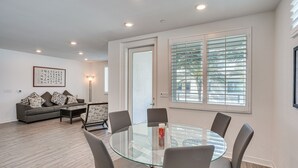 A modern living room with a glass-top dining table, grey sofas, and a large window with blinds. - Convenient Anaheim Condo: Walk to Angel Stadium (Anaheim)