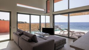 A modern living room with a large sofa, a flat-screen TV, and a dining table. There is a balcony with a view of the ocean and a pool. - Casa Tarrha - Beachside Retreat with Private Heated Pool (Chania Town  Livadia)