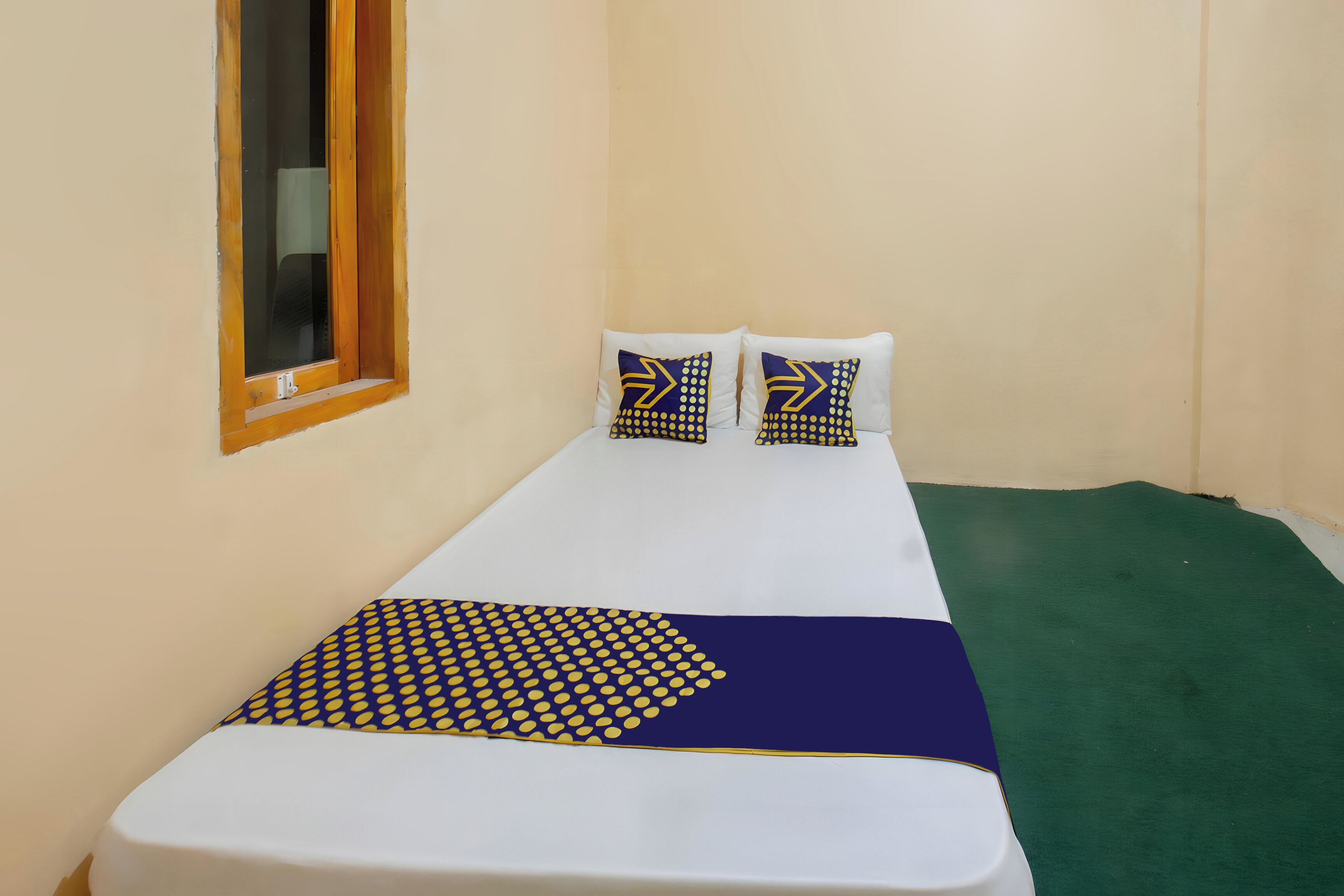 Una cama individual con colchón blanco, dos almohadas con diseño geométrico, una colcha azul y amarilla, y una alfombra verde.