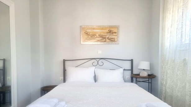 Una cama bien hecha con sábanas blancas y dos toallas dobladas al lado, una mesita de noche con una lámpara, y una obra enmarcada sobre la cama.