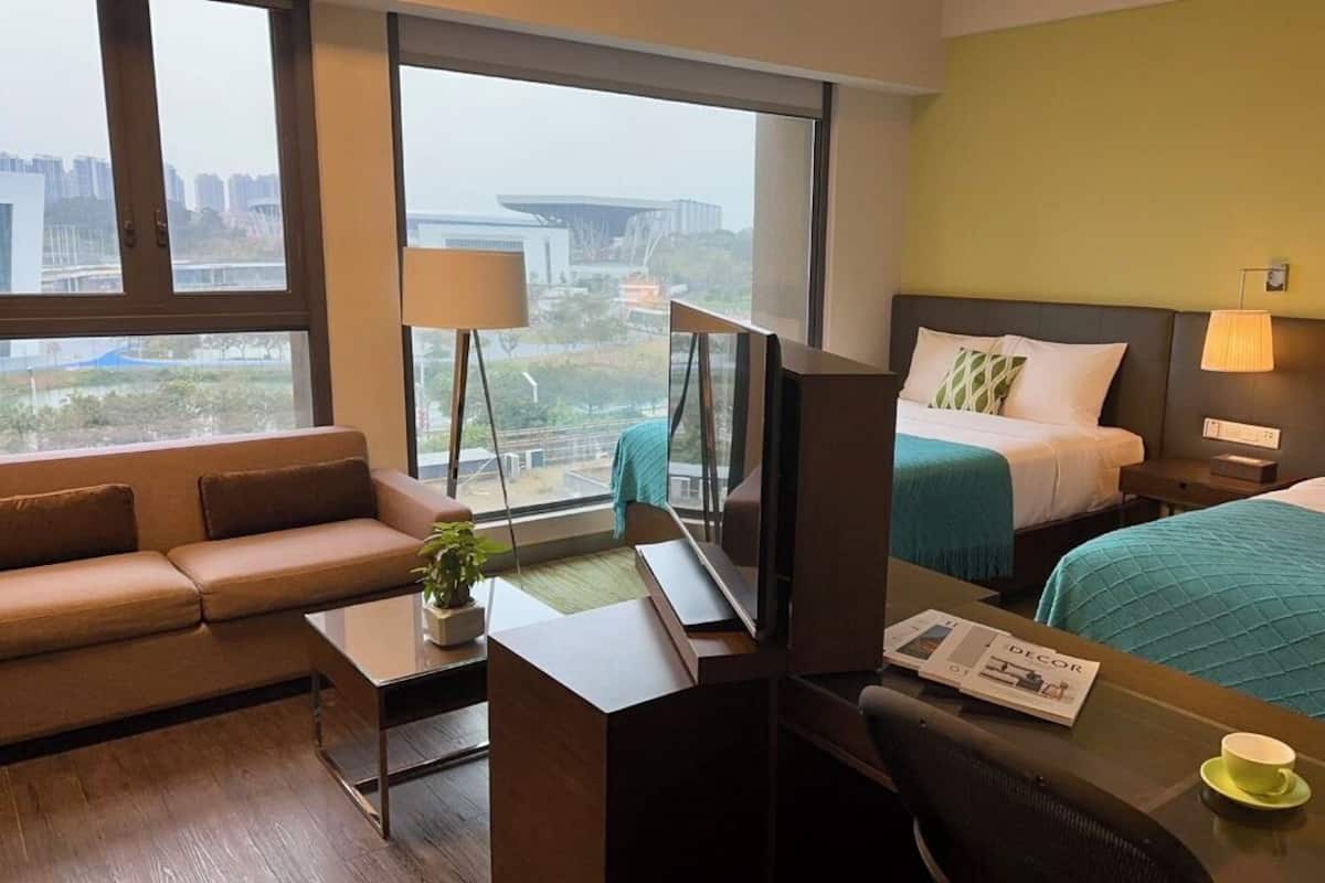 A hotel room with a sofa, a desk with a laptop, a bed with a green blanket, and a view of the cityscape.