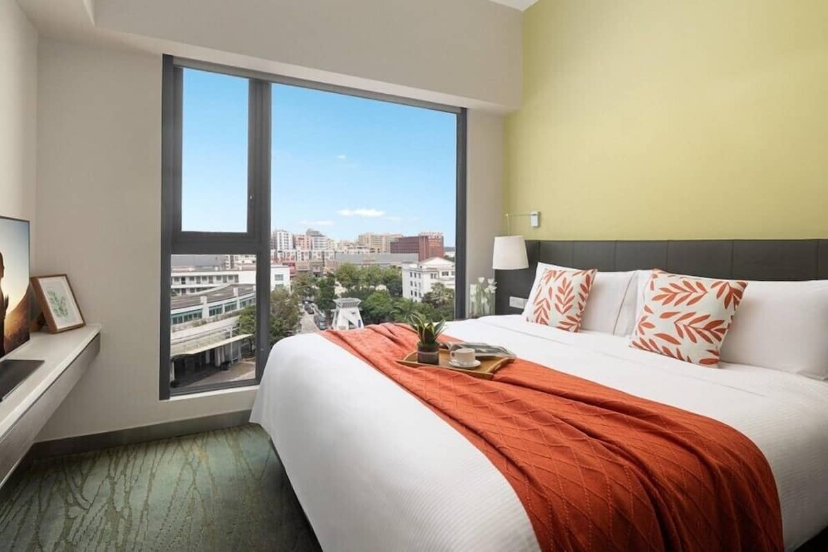 A hotel room with a large window, a bed with white and orange bedding, a bedside table with a lamp, and a view of the cityscape.