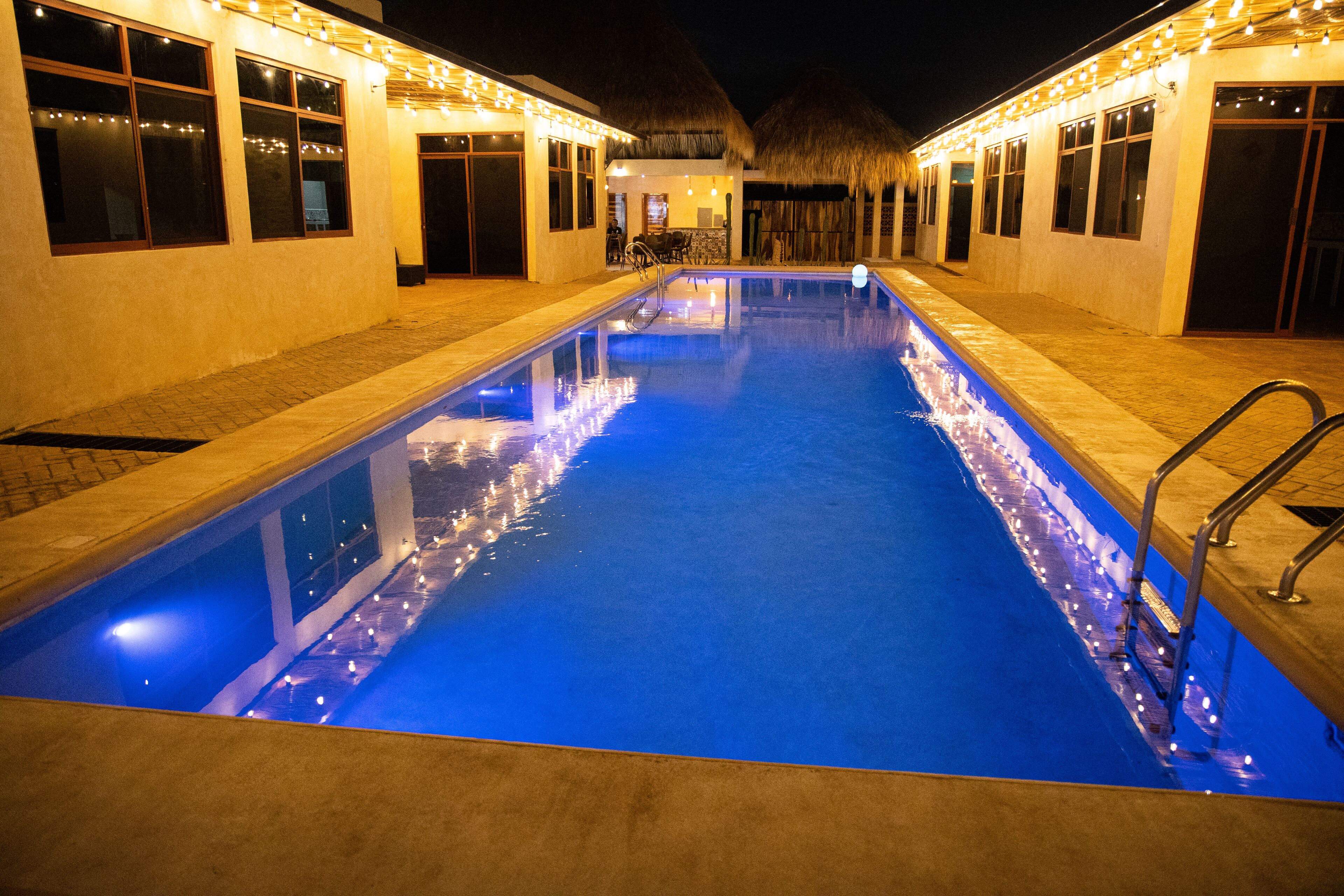 A swimming pool with a blue tint, surrounded by a building with multiple windows and a thatched roof structure.