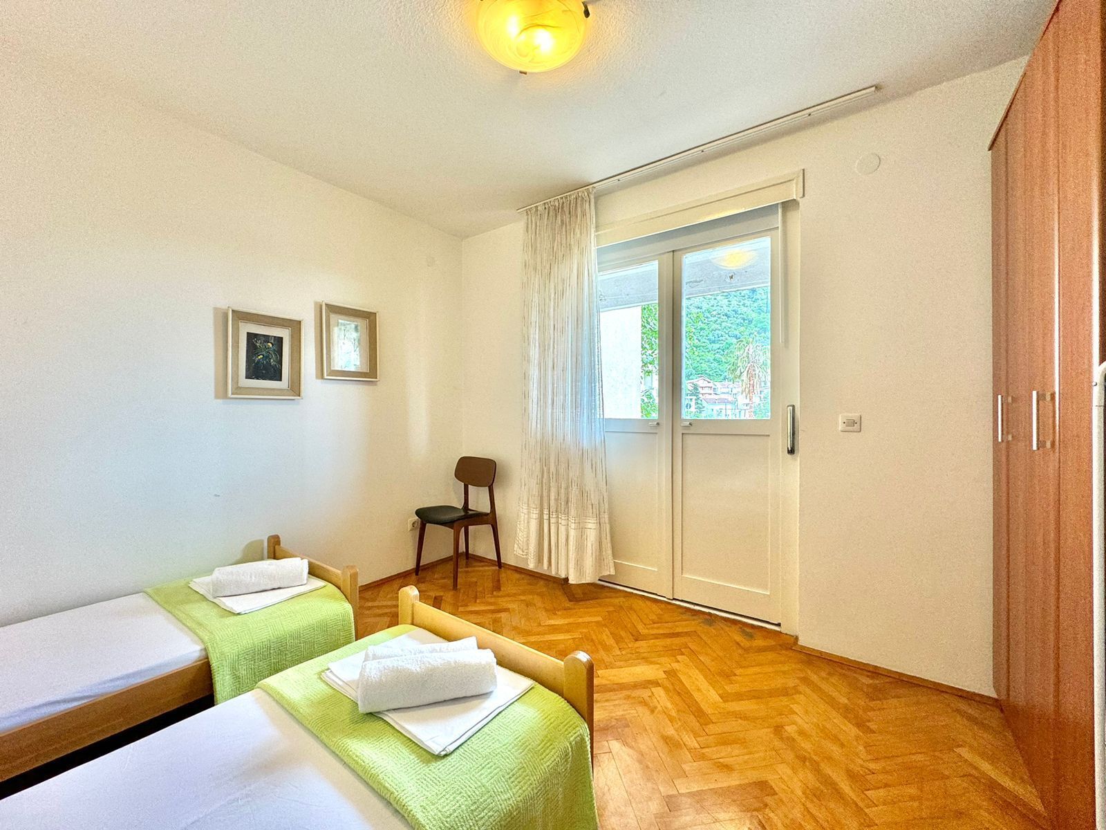 A room with two beds, a chair, a window with curtains, and a wooden floor.
