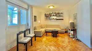 A living room with a sofa, two chairs, a coffee table, and a TV stand with a TV. There are shelves above the sofa with various items. - Family two bedroom apartment Slavia (Budva)