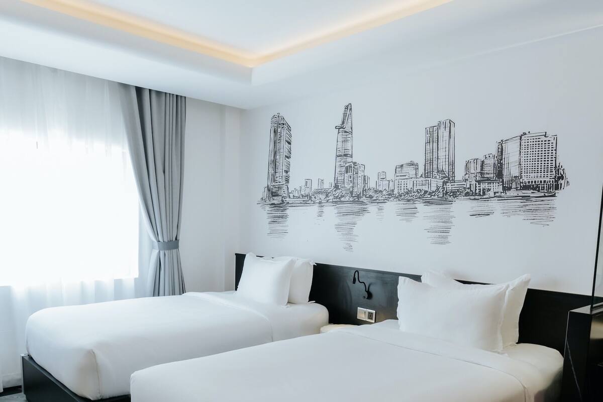 A hotel room with two beds, a cityscape wall art, a large window with curtains, and a wooden floor.