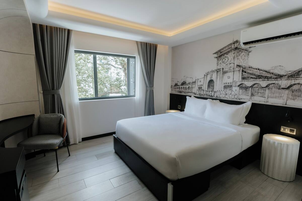 A modern hotel room with a large bed, a chair, a bedside table, and a window with curtains.