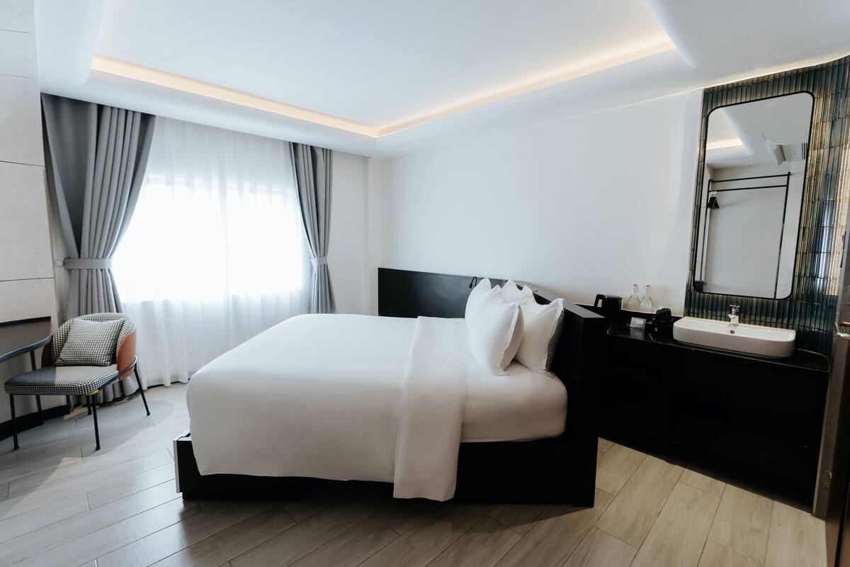 A modern hotel room with a large bed, a chair, a bathroom with a mirror, and a window with curtains.