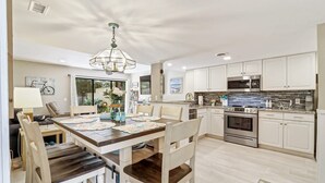 A modern kitchen with white cabinets, stainless steel appliances, and a dining area with a wooden table and chairs. - Beach Club Bliss Villa Steps to Sand, Shops & Sunshine (Hilton Head Island)