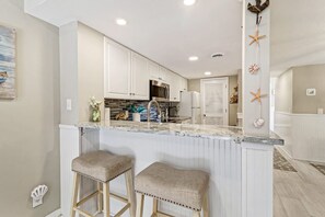 A kitchen with white cabinets, a granite countertop, two bar stools, a refrigerator, and a microwave.