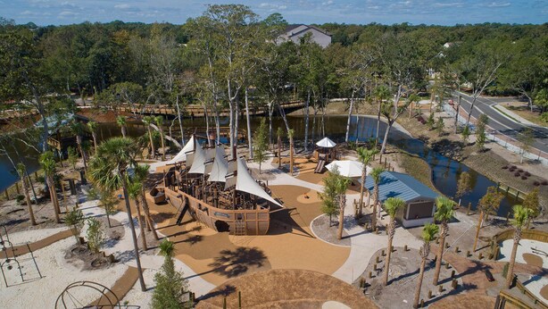 A resort area with a sandy beach, palm trees, a water body, and a wooden structure with a sail-like roof. - Beach Club Bliss Villa Steps to Sand, Shops & Sunshine (Hilton Head Island)