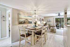 Un coin repas avec une table en bois, des chaises et un lustre. Un tableau orne le mur et une porte vitrée offre une vue sur l’extérieur.