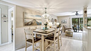 A dining area with a wooden table, chairs, and a chandelier. There is a painting on the wall and a view of the outdoors through a glass door. - Beach Club Bliss Villa Steps to Sand, Shops & Sunshine (Hilton Head Island)