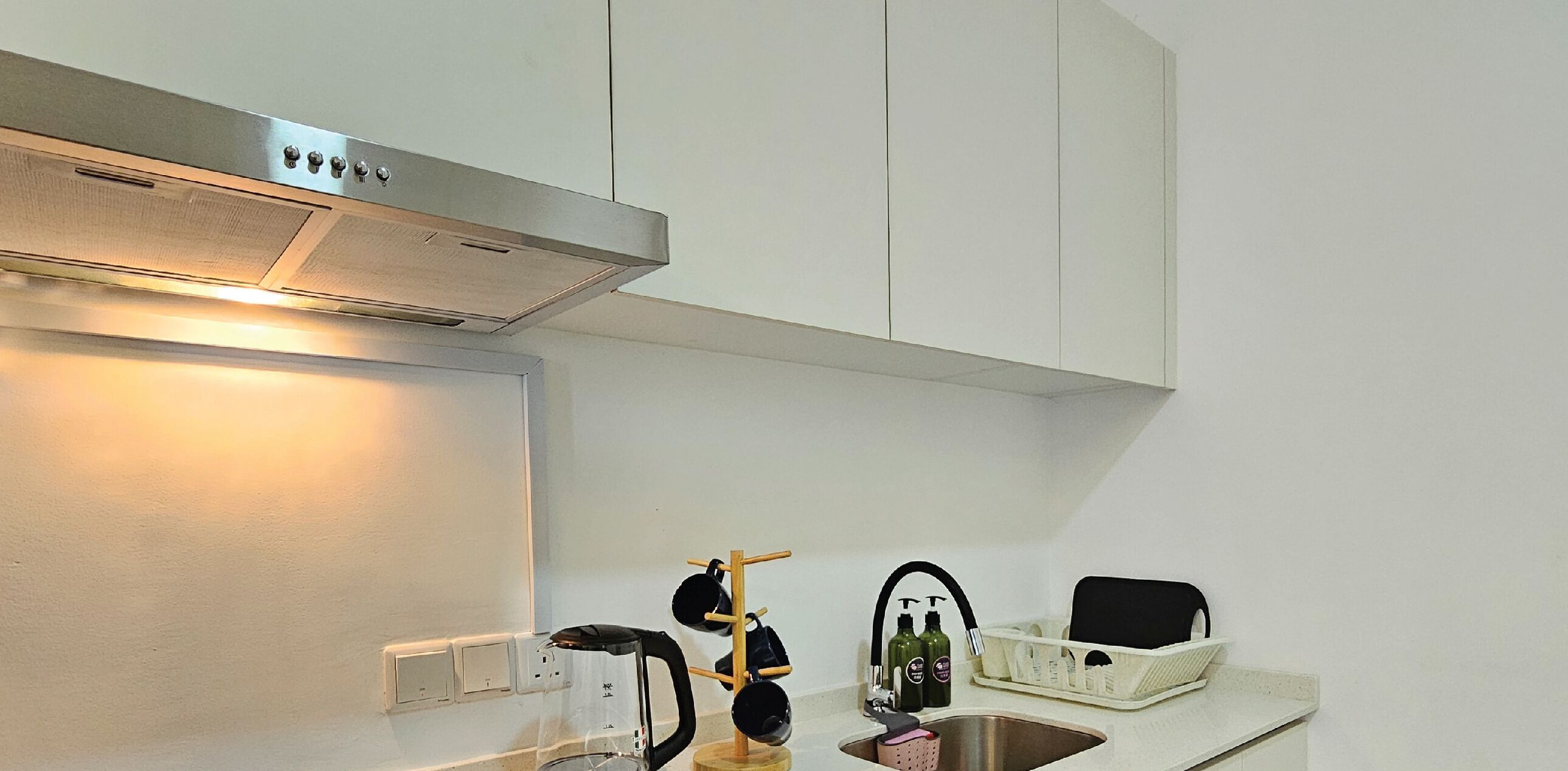 A kitchen with white cabinets, a stainless steel range hood, a coffee maker, a kettle, a drying rack, and a sink.