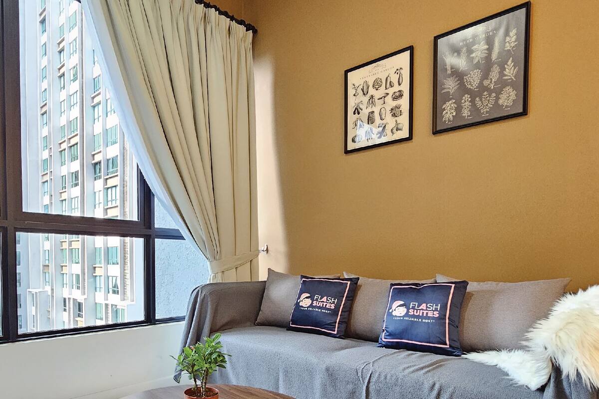 A modern hotel room with a large window, a grey sofa with branded pillows, and two framed botanical prints on the wall.