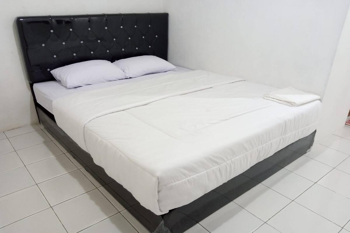 Una cama bien hecha con cabecera negra y ropa de cama blanca en una habitación con suelo de baldosas blancas.