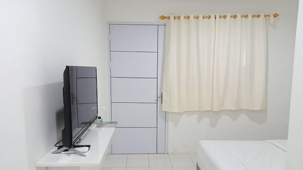 Una habitación pequeña y limpia con una cama, un televisor sobre un soporte, una puerta blanca y una cortina.