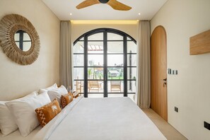 A bedroom with a large bed, a ceiling fan, a round mirror, and a view of the outdoors through a large window. - Villa The Santé (Canggu)