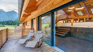 Une terrasse en bois avec des chaises longues, une porte vitrée donnant sur un balcon doté d’une balustrade en bois, et une vue sur les montagnes en arrière-plan.