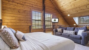 Une pièce chaleureuse avec un plafond en bois, un lit recouvert de linge de maison blanc, un canapé gris agrémenté de coussins à motifs et un téléviseur fixé au mur.