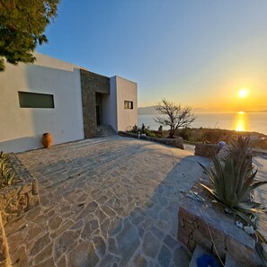A modern building with a stone pathway leading to a door, a large window, and a view of the sunset over water.