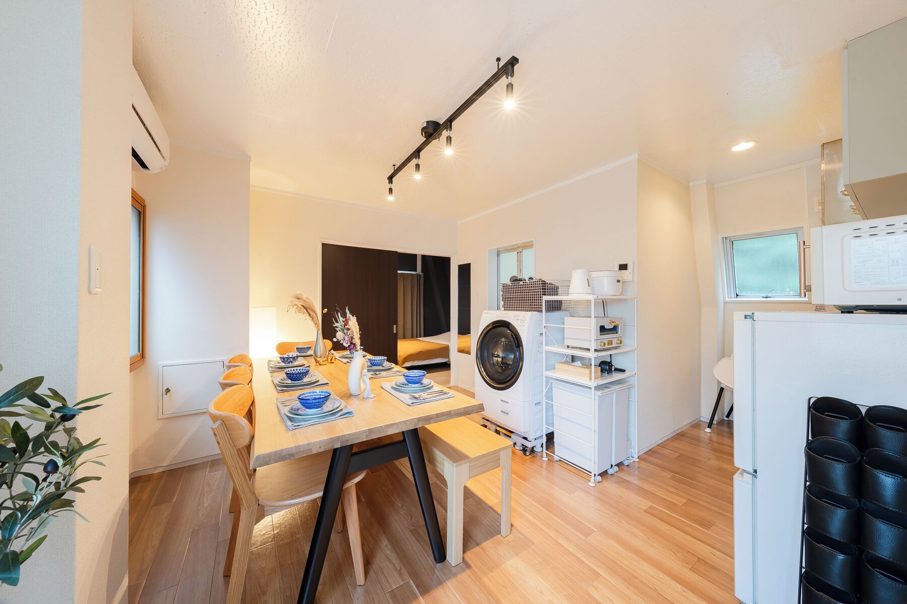 A compact kitchen with a washing machine, a dining area with a table set for two, and a small seating area with a bench.
