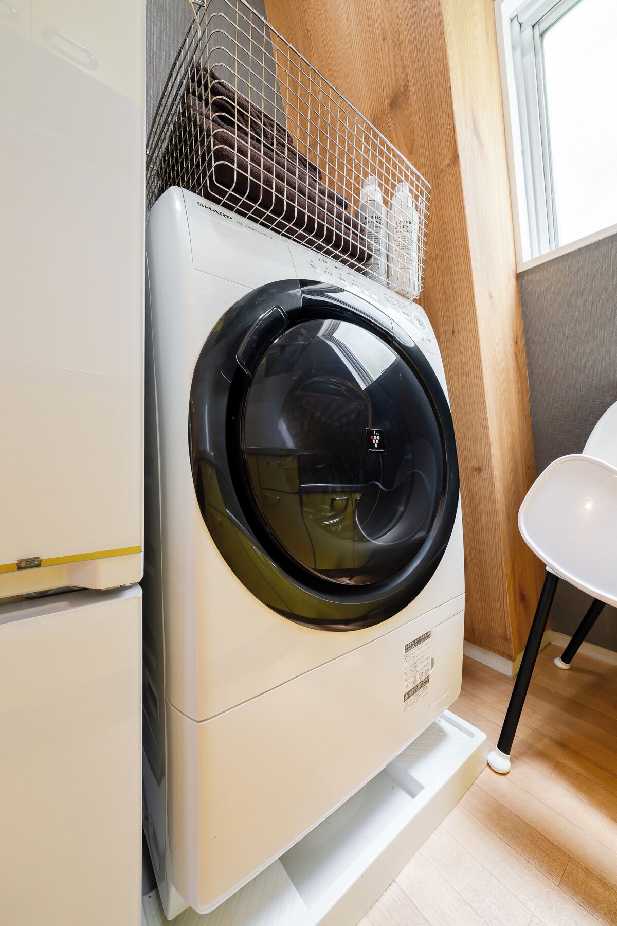 A front-loading washing machine with a basket on top, placed in a kitchen with wooden cabinets and a white chair.