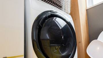 A front-loading washing machine with a basket on top, placed in a kitchen with wooden cabinets and a white chair.