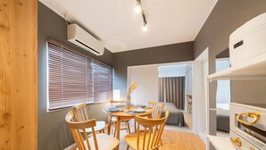 A compact room with a dining table, wooden chairs, a white shelf with a golden handle, and a window with blinds.