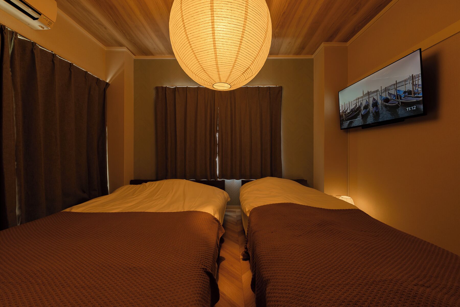 A hotel room with two beds, a large hanging light fixture, a television on the wall, and a window with curtains.