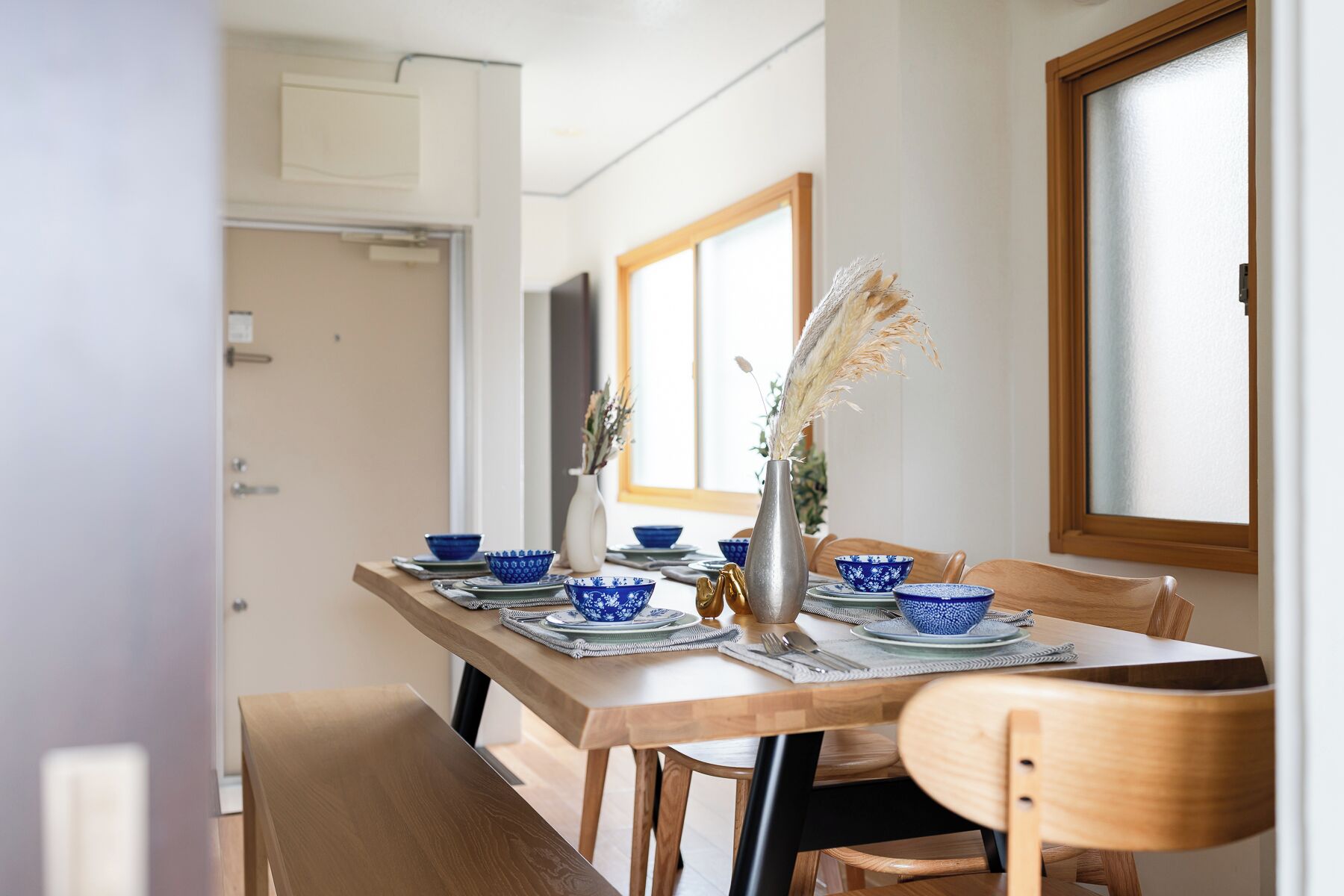 A dining table set with blue patterned bowls, a silver vase with pampas grass, and wooden chairs.