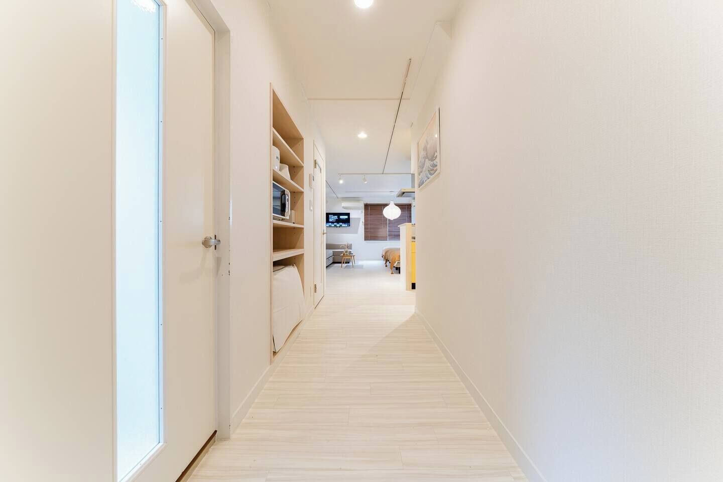 A narrow hallway with a wooden floor, white walls, and a door on the left. Shelves with various items are visible on the right.