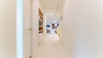 A narrow hallway with a wooden floor, white walls, and a door on the left. Shelves with various items are visible on the right.