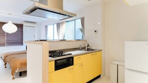 A compact kitchen with a gas stove, sink, and a small refrigerator. There is a window with curtains and a bed with a wooden headboard in the adjacent room.