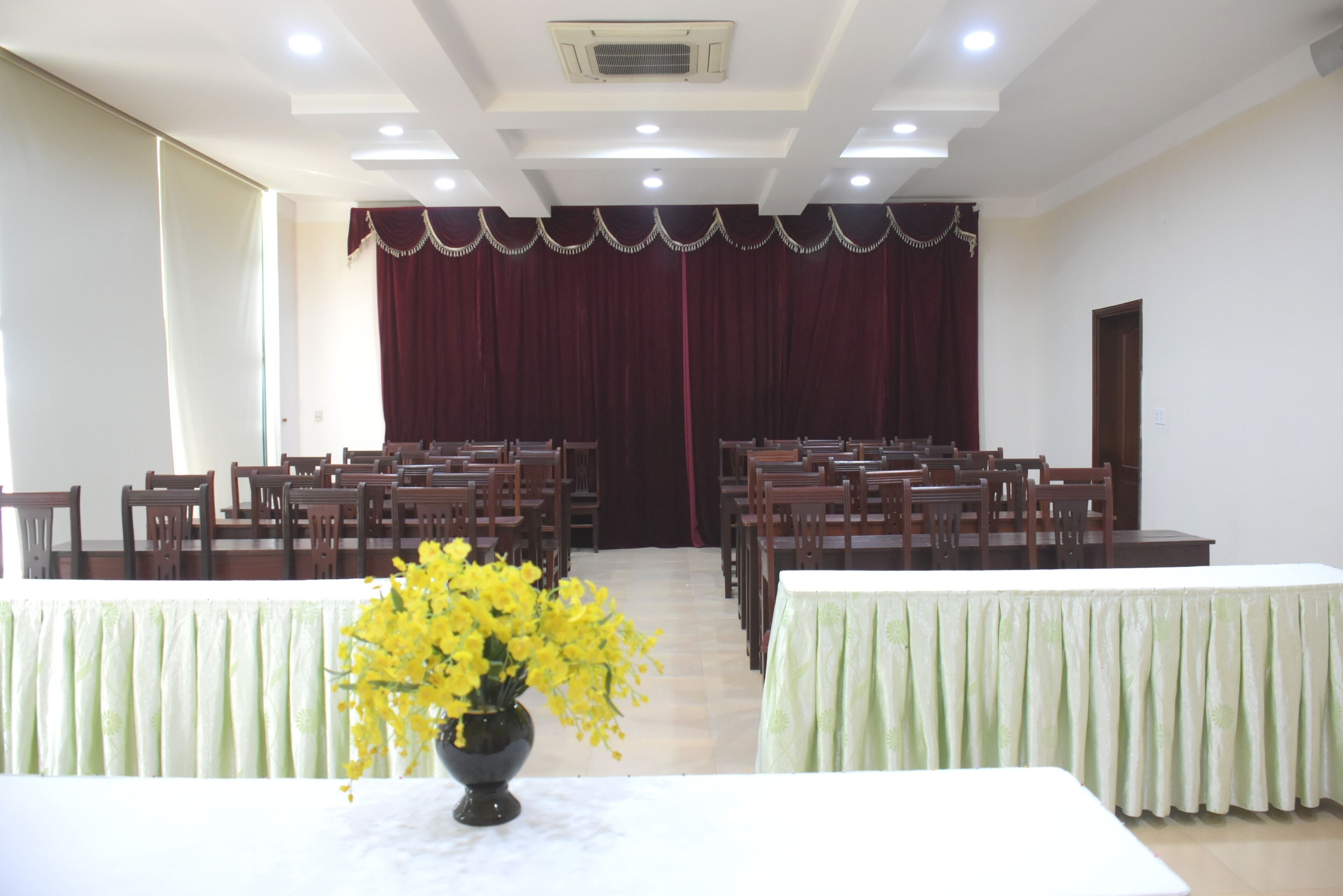A conference room with rows of wooden chairs and tables, a stage with a red curtain, and a vase of yellow flowers on the table.
