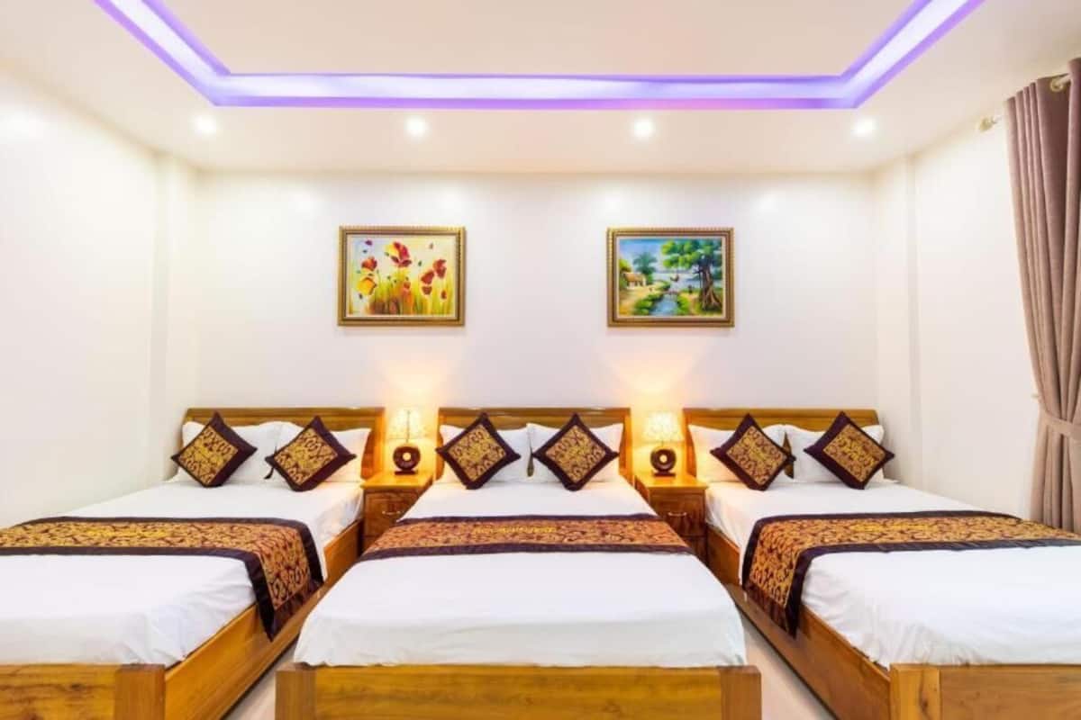 Habitació d'hotel amb tres llits de fusta, cadascun amb roba de llit blanca i almoinades decoratives. Hi ha dos quadres penjats a la pared, un amb flors i l'altre amb un paisatge. La habitació té un sostre amb il·luminació morada.