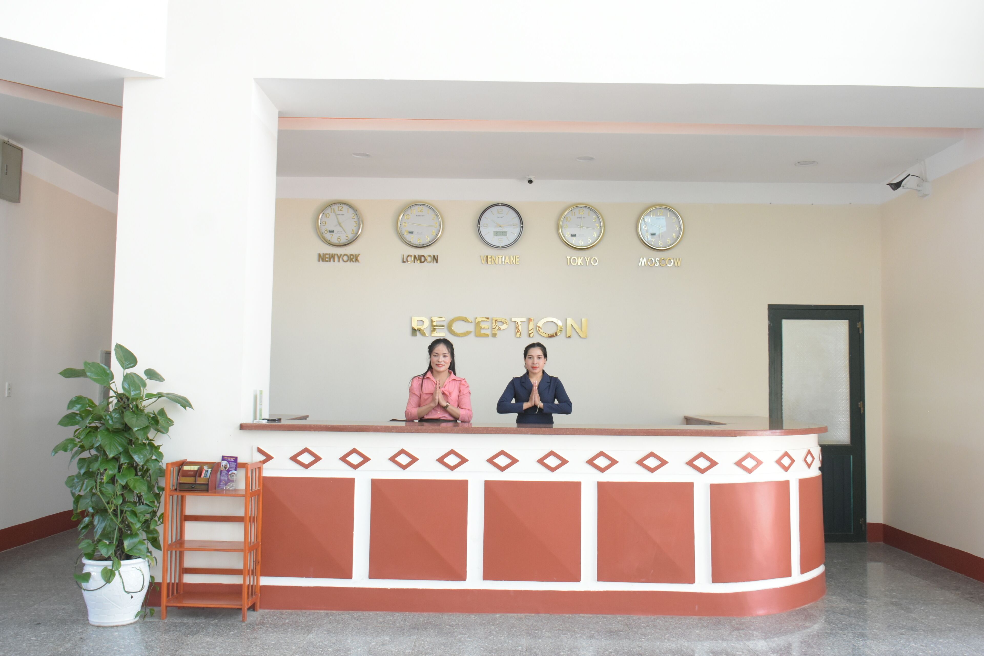 Un mostrador de recepción con dos empleados, una planta en maceta y relojes que muestran horarios de diferentes ciudades.