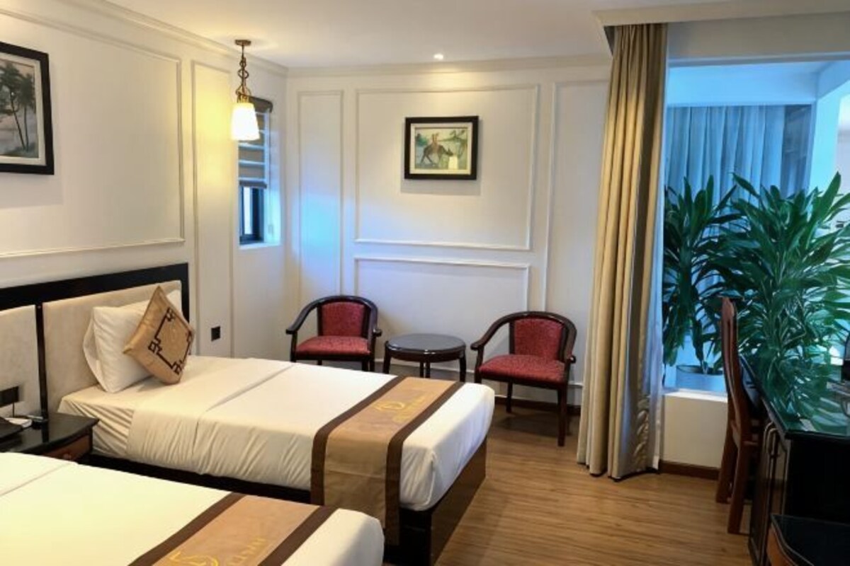 Ett hotellrum med två sängar, ett skrivbord, två stolar, ett litet bord, en växt och ett fönster med gardiner.