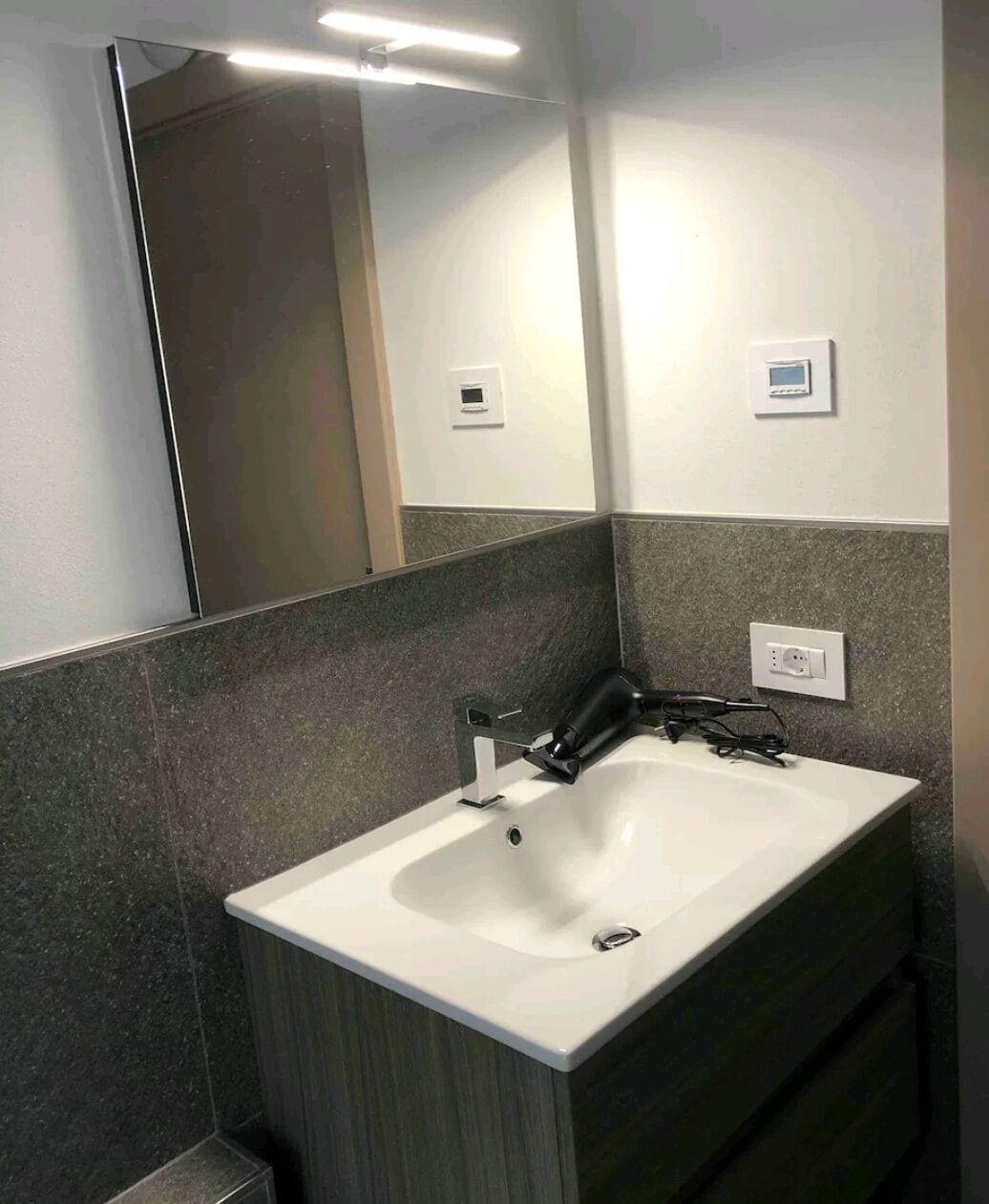 Une salle de bain avec un lavabo blanc, des murs recouverts de carreaux foncés, un miroir et un interrupteur.