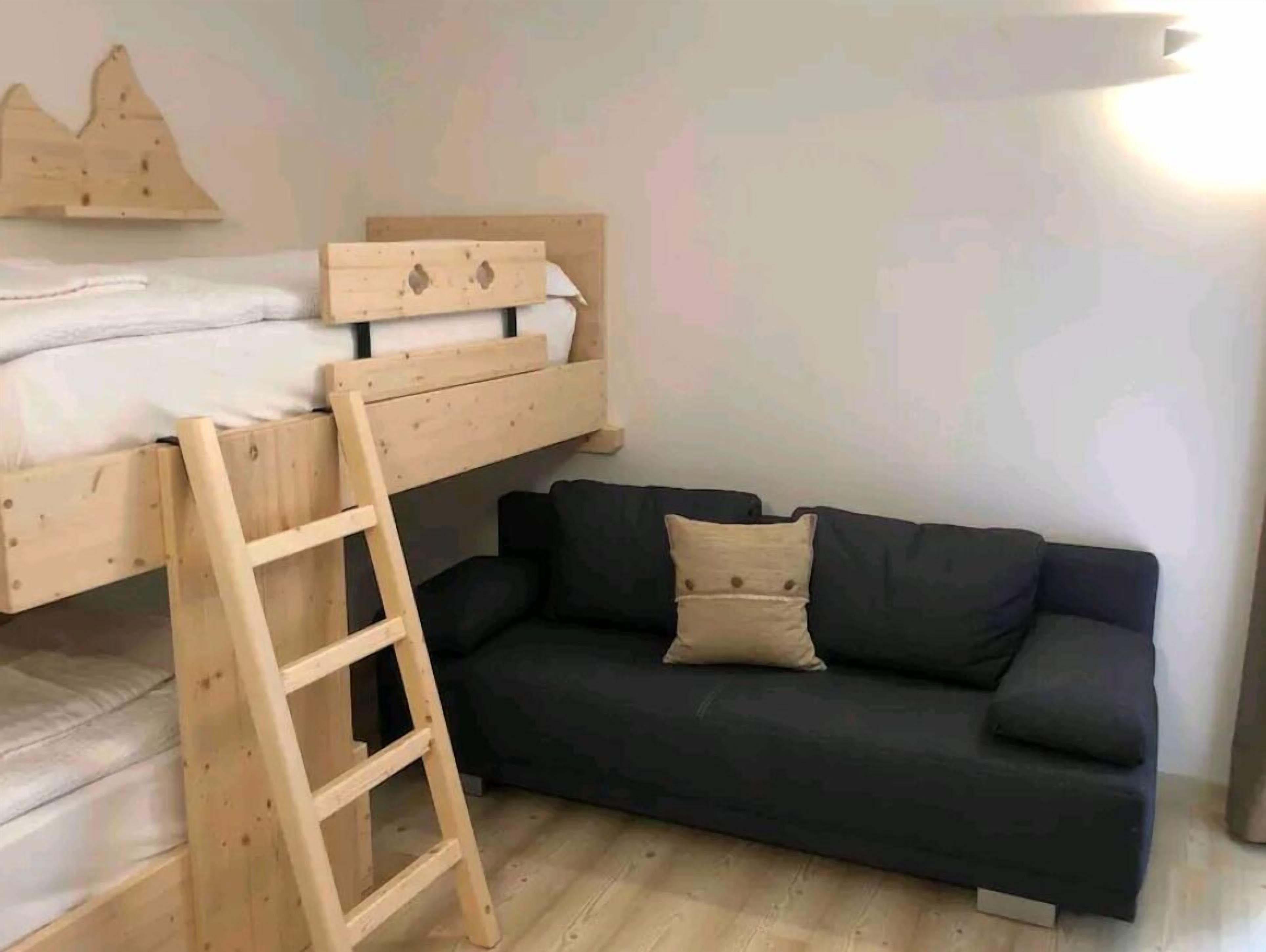 A room with a bunk bed, a wooden ladder, and a dark-colored sofa with cushions.
