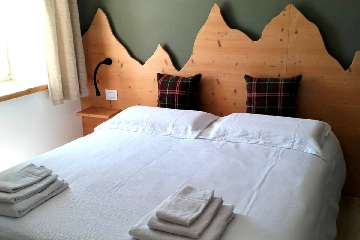 A neatly made bed with white linens and folded towels, a wooden headboard with a unique shape, and a nightstand with a lamp.